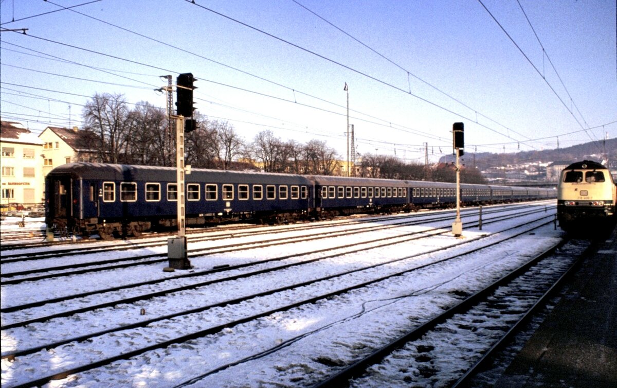218 406-7 (in türkis-beige; aktuell in verkehrsrot mit Lätzchen in Kempten im Einsatz) und links eine lange Reihe blauer Personenwagen Bm (2.Klasse) in Ulm am 30.01.1981.