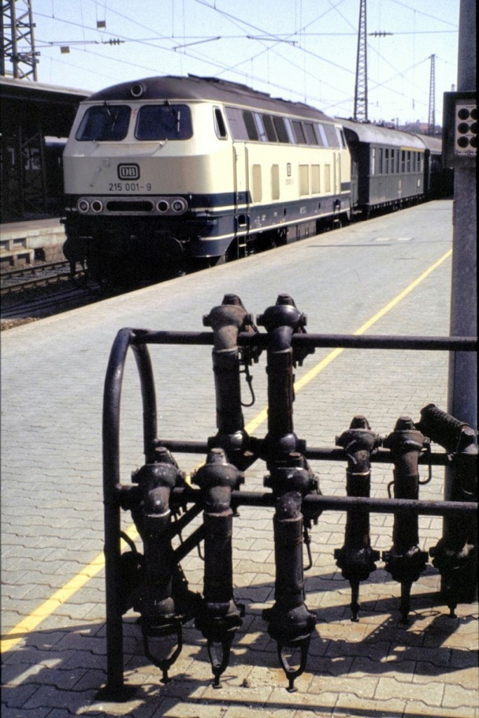 Das bunte Leben der 215 001-9, in ozeanblau/beige in Ulm am 09. Juli 1982. Im Vordergrund sind noch die Verbindungsschläuche für die Dampfheizung der alten Reisezugwagen zu sehen; im Juli gibt es jedoch keine Verwendung. Der erste angehängte Wagen ist ein AByse 633. (Diascan)