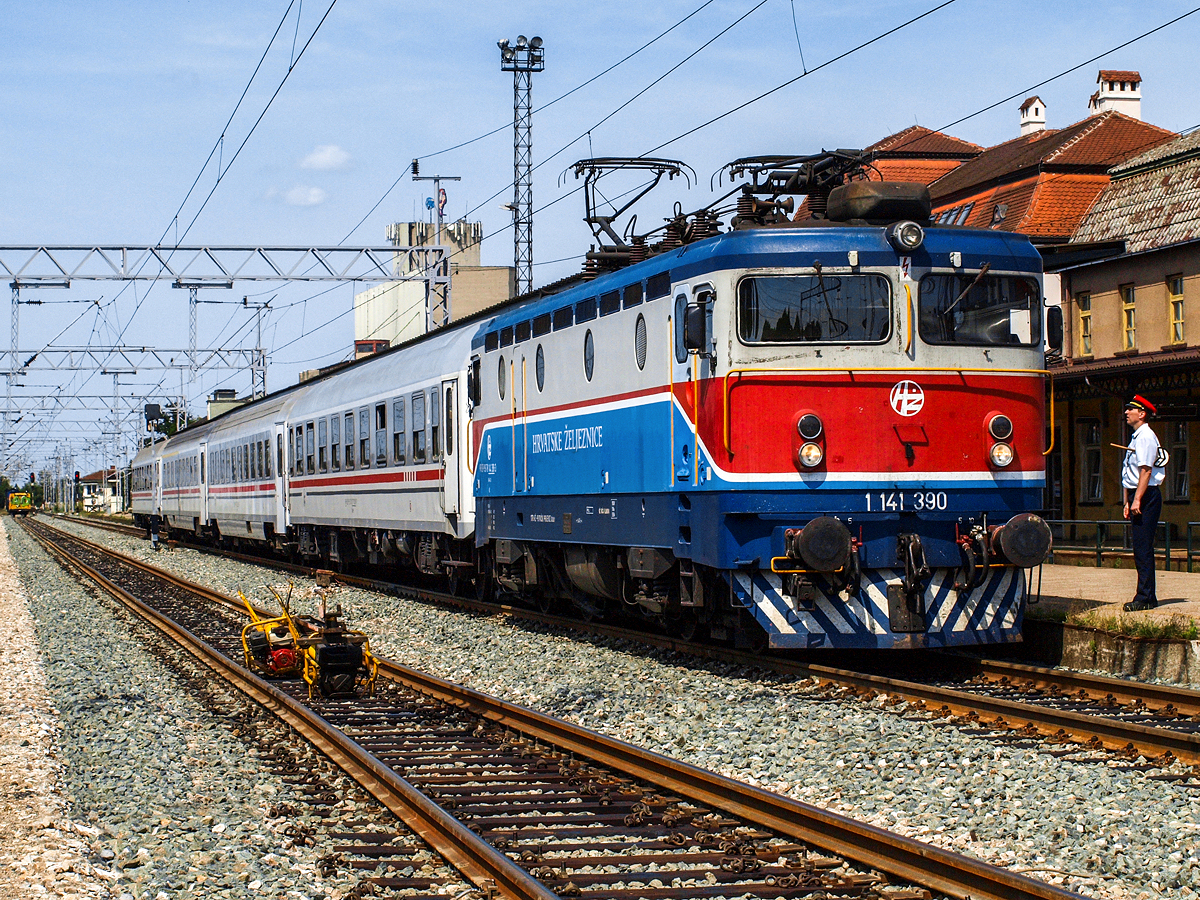 In Bahnknoten Koprivnica im Norden Kroatiens werden die Fernverkehrszüge von Osijek nach Zagreb auf elektrische Traktion umgespannt, dazu ist ein Aufenthalt von 10 bis 15 Minuten vorgesehen. Tief im kroatischen Hinterland sind die Somemrtage üblicherweise ausgesprochen heiß, so auch am frühen Nachmittag des 30.06.2015. Die 1141 390 hat gerade den Schnellzug 702 von Osijek nach Rijeka übernommen und wartet auf den Abfahrtsbefehl, um die Fahrt in Richtung Zagreb fortzusetzen.