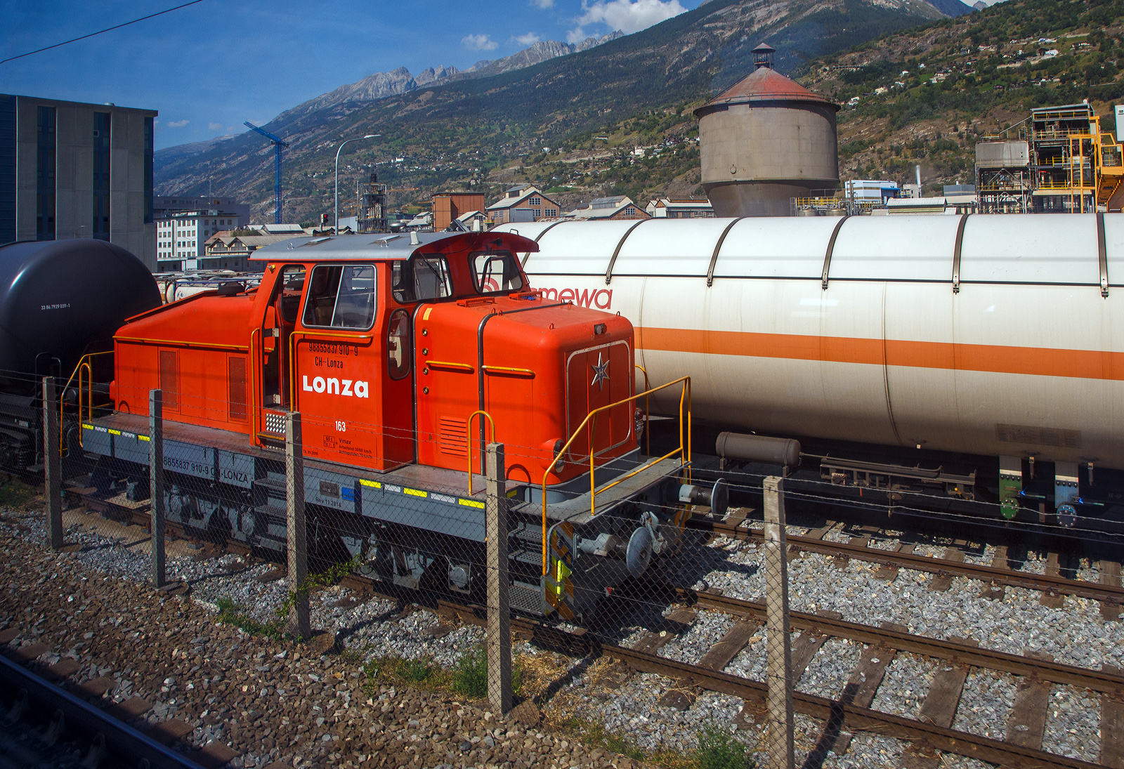 Aus dem Zug heraus, die Em 3/3 163  Rene  (98 85 5837 910-9 CH-LONZA) eine Henschel DHG 500 C der Lonza  AG (Visp), steht am 08.09.2021 in Visp.

Die Henschel  DHG 500 C wurde 1968 von Henschel in Kassel unter Fabriknummer  31239  gebaut und war erst als Mietlok im Einsatz. Im Jahr 1971 verkaufte sie dann Henschel an die Lonza AG in die Schweiz. 2014 wurde die Lok von der LSB Lok Service Balmer AG in Hinwil komplett neu revidiert und remotorisiert.

Die Henschel DHG 500 C ist eine dieselhydraulische Lokomotive, die von den Henschel-Werken gebaut wurde. Sie war vor allem für den Einsatz im schweren Werksdienst vorgesehen. Die Lok gehört zur ab 1962/63 angebotenen sogenannten vierten Generation der Henschel-Loks, der ersten Generation mit Gelenkwellenantrieb. Die Aufbauten wurden dabei ohne große Änderungen von der vorigen Generation übernommen.  Zur besseren Bogenläufigkeit war die mittlere Achse seitenverschiebbar gelagert. 

Von den Henschel DHG 500 C wurde zwischen 1963 und 1976 insgesamt  62 Exemplaren gebaut. Die Loks gingen vornehmlich an Bergbaugesellschaften und Stahlproduzenten sowie Chemieunternehmen. So erwarb die Rheinelbe Bergbau AG in Gelsenkirchen elf Loks und die Hibernia AG, Zechenbahn- und Hafenbetriebe Ruhr-Mitte in Gladbeck neun Loks vom Typ DHG 500 C. Die meisten Lokomotiven wiesen bei Auslieferung den Henschel-Standardanstrich in Blau mit zwei horizontalen Zierstreifen an den Vorbauten auf. Die durch Zusammenschluss entstandene Ruhrkohle AG hatte zeitweise 31 Lokomotiven dieses Typs im Bestand. 

TECHNISCHE DATEN (nach Umbau):
Spurweite:  1.435 mm (Normalspur)
Achsformel: C
Länge über Puffer:  9.900 mm
Höhe:  4.225 mm
Breite: 3.040 mm
Fester Radstand:  3.700 mm
Kleinster bef. Halbmesser:  80 m
Dienstgewicht:  60 t
Höchstgeschwindigkeit: 30 km/h / 60 km/h (Schleppfahrt 70 km/h)
Treibraddurchmesser:  1.000 mm
Motor:  neuer Deutz V6 Motor mit elastischer Vorschaltkupplung. inkl. Partikelfilter (Vollstrombrenner mit aktiver Regeneration), 
der ehemalige Motor war ein Henschel 12V 1416 
Installierte Leistung:  368 kW (500 PS)
Getriebe:  Voith
Leistungsübertragung:  hydraulisch (über Gelenkwellen)
