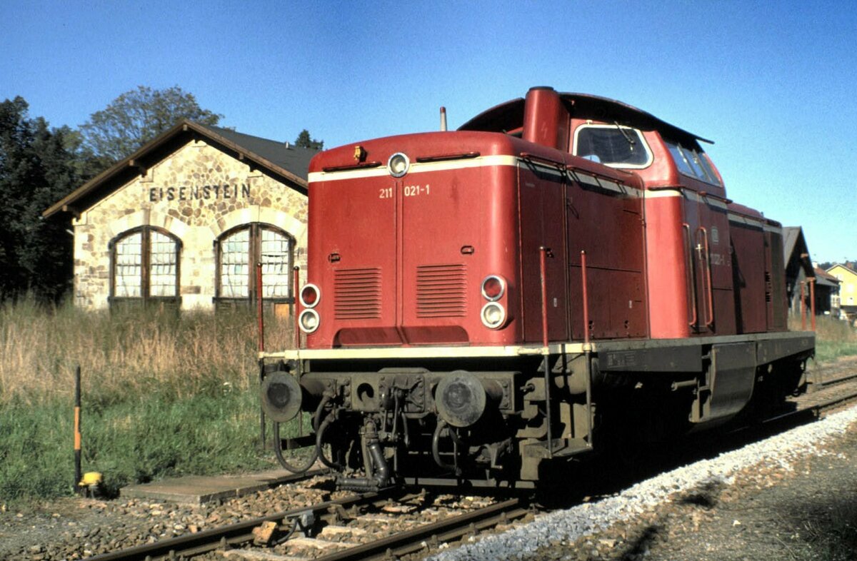 211 021-1 vor dem Lokschuppen in Bayerisch Eisenstein im Oktober 1989. Im Lokschuppen werden zu diesem Zeitpunkt bereits Exponate für das Bayerische Eisenbahnmuseum gesammelt.