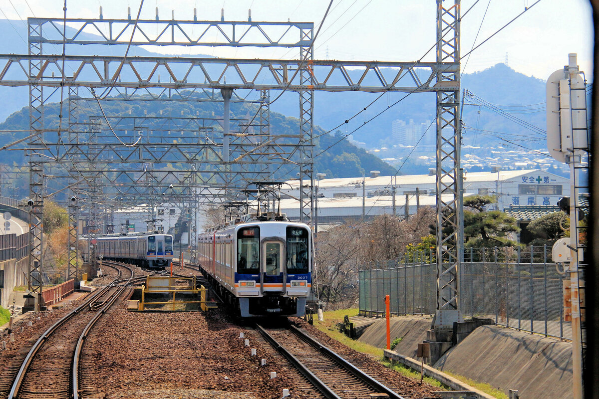 Der Nankai-Konzern, Partnerbahn der MOB: Die Kôya-Linie des Nankai-Konzerns führt von Osaka (3, 4 m.ü.M.) aus zuerst durch die südlichen Vororte der Stadt und steigt dann durchs Gebirge im südlichen Hinterland - wo auch schon städtische Wohnblöcke in prachtvoller Natur stehen - bis zum Scheiteltunnel auf der Passhöhe von Kimi Tôge (248, 2 m.ü.M.). Von da  stürzt  die Linie auf einer langen und kurvenreichen Steilrampe ins Tal des Kinokawa Flusses hinunter, zum Städtchen Hashimoto (90, 6 m.ü.M.). In Hashimoto beginnt dann die eigentliche Bergstrecke zum Kôya-san. Im Bild zwei Züge des Nankai-Konzerns in Mikkaichi-chô (Nr. 2037 und 1001), unterwegs zwischen Osaka und dem Kimi Tôge-Pass. Im Hintergrund sieht man die sich weit in diese bäuerliche Gegend hineinfressenden Ueberbauungen für  Natursuchende  aus der Riesenstadt Osaka. 28.März 2017  