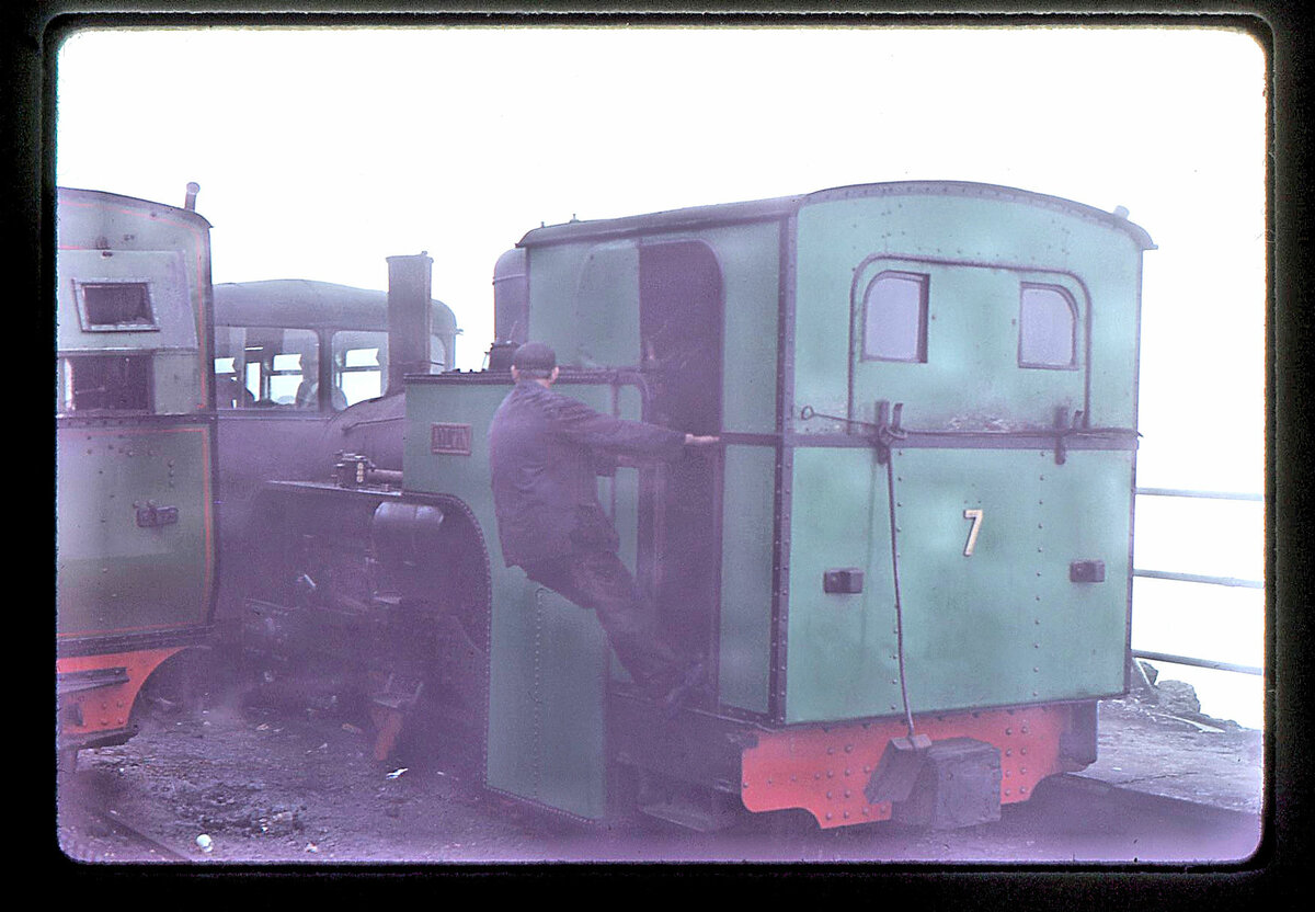 Die in der Schweiz gebauten Lokomotiven der Snowdon Mountain Railway / Rheilffordd yr Wyddfa - Lok 7. Leider wurden die beiden neusten Dampfloks (gebaut von SLM 1923) 1990 (Lok 7) und 1992 (Lok 8) ausgemustert und zerlegt. Ein Wiederaufbau ist aus Kostengründen nicht geplant. Das ist besonders schade, wenn man bedenkt, dass keine solche schweizerische Lok in der Schweiz selbst noch vorhanden ist. Die Loks haben einen Ueberhitzer und grössere Wassertanks als Lok 6. Im Bild steht Lok 7 im Nebel der Bergstation, 21.Juli 1974. Die Lok trug hier den Namen  Aylwin  (eine literarische Romanze von 1898).  