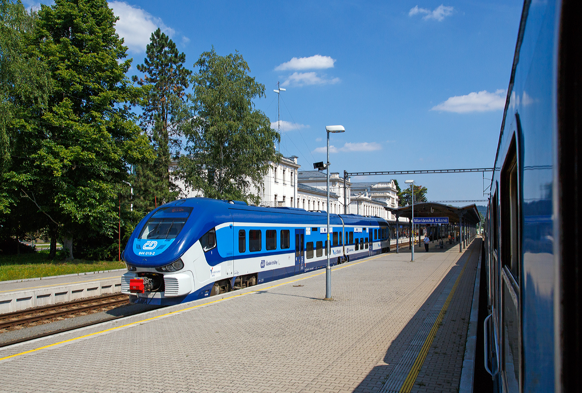 Ein letzter Blick aus dem Zug (Rx 760  Vyehrad ) heraus auf den Bahnhof Marinsk Lzně (deutsch Marienbad) am 01.07.2015. 

Auf Gleis 2 steht der „RegioShark“  ČD 844 018-2 (CZ ČD 95 54 5 844 018-2), ein Dieseltriebzug vom Typ PESA LINK II, als Os 7382 zur Abfahrt bereit. Er hatte mich zuvor nach Marienbad gebracht.