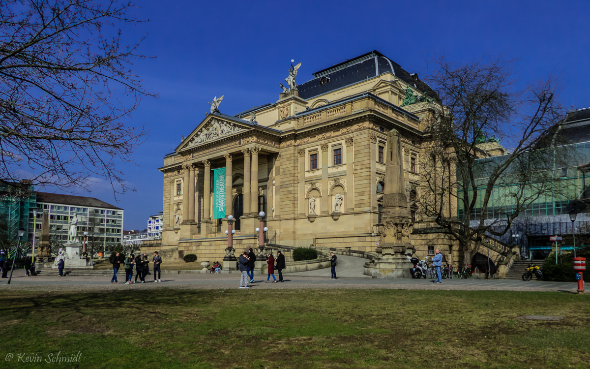 Hier ist die Schauseite des Hessischen Staatstheaters Wiesbaden zu sehen. Der eigentliche Haupteingang befindet sich direkt auf der anderen Seite in den Theaterkolonnaden. Es handelt sich um ein neobarockes Bauwerk der Wiener Theaterarchitekten Ferdinand Fellner d. J. und Hermann Helmer von 1892 bis 1894. Rechts befindet sich ein architektonisch kontrastierender Werkstätten-Anbau aus den 1970er-Jahren. (25.03.2018)