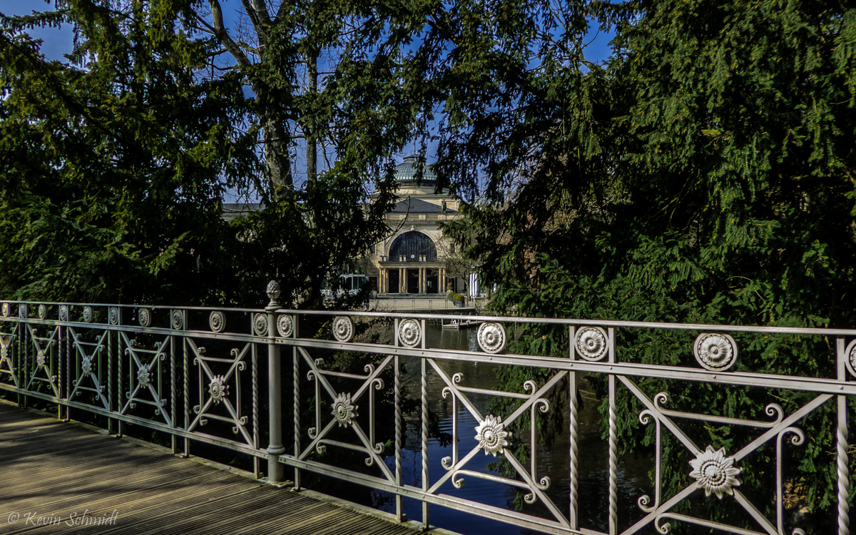 Im <u><a href= https://www.wiesbaden.de/leben-in-wiesbaden/freizeit/natur-erleben/gruenanlagen-parks/kurpark-wiesbaden.php  target= _blank >Wiesbadener Kurpark</a></u> führt diese Brücke mit einem hübschen Geländer auf eine kleine Halbinsel im Kurpark-Weiher, von wo aus man die Rückseite des Kurhauses zwischen den Bäumen hindurch erspähen kann. (25.03.2018)