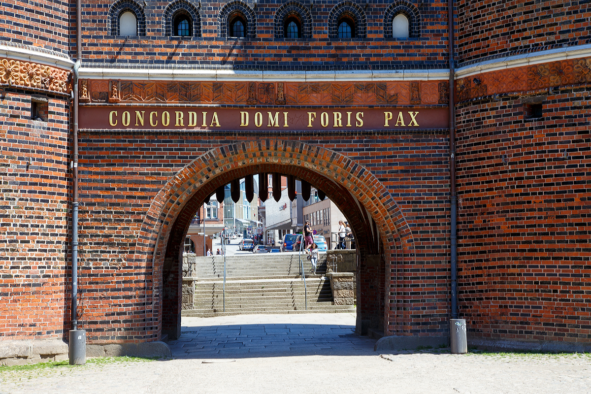 
Lübeck am 11.06.2015, Deteil vom Holstentor von Westen (Feldseite) mit der Innschift: 
Concordia domi foris pax („Eintracht innen, draußen Friede“). 

Dieser Schriftzug stammt von 1871 und ist eine verkürzte Form der Inschrift, die zuvor auf dem (nicht erhaltenen) Vortor gestanden hatte: Concordia domi et foris pax sane res est omnium pulcherrima („Eintracht innen und Friede draußen sind in der Tat für alle am besten“).

Obwohl mir die Übersetzung der heutigen Inschrift mit Google besser gefallen würde, hier würde es wie folgt übersetzt: Eintracht und Frieden im In- und Ausland (spricht auf der Welt)
