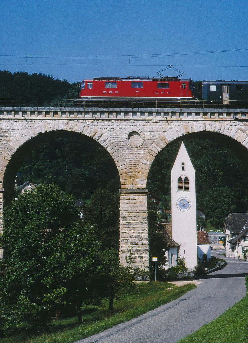 SBB: Obschon in der Schweiz jeden Tag sehr viele Re 4/4 mit Reise- Güter- und Postzügen unterwegs sind, handelt es sich hier um eine sehr seltene Aufnahme. Zur Abwechslung konnte im Sommer 1996 eine Re 4/4 I der zweiten Serie mit einem umgeleiteten Reisezug vor der herrlichen Kulisse der Kirche Läufelfingen beim Passieren des Viadukts verewigt werden.
Foto: Walter Ruetsch  