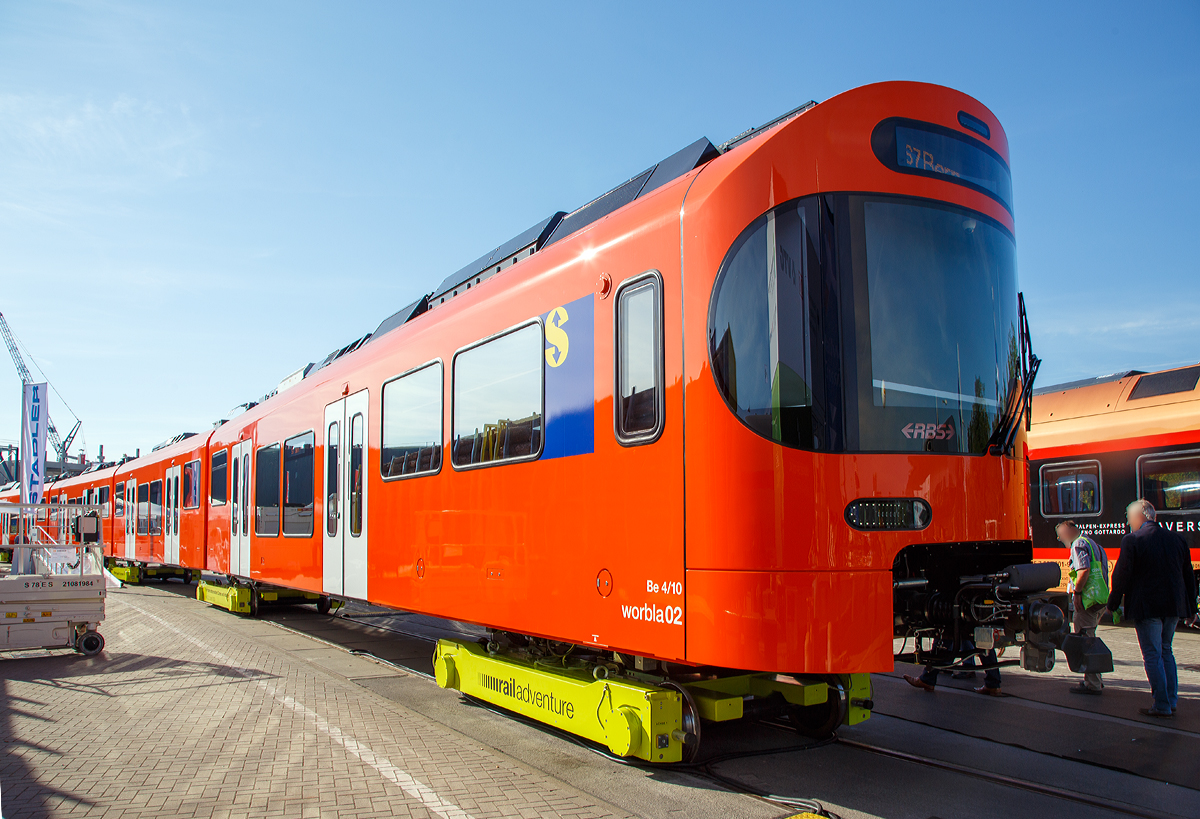 Stadler Rail präsentierte auf der InnoTrans 2018 (hier 18.09.2018) den neuen Worbla-Zug Be 4/10 für die RBS (Regionalverkehr Bern-Solothurn), hier in Form von dem RBS Be 4/10 Worbla 02. Bis 2020 sollen alle Mandarinli durch die Worblas bei der RBS ersetzt werden.
Der Triebzug steht hier auf Rollböcke der RailAdventure GmbH („Loco Buggies“).

RBS hat im Juni 2016 bei Stadler in Bussnang 14 vierteilige S-Bahn-Züge vom Typ Be 4/10 Worbla (Elektrischer Niederflurtriebzug) für die Linie S7 bestellt. Die vierteiligen elektrischen Meterspurzüge weisen 2 Triebköpfe am Ende und 3 Jakobs-Laufdrehgestelle als Verbindung zwischen den 4 Wagenkästen auf. Die Züge sind für eine maximale Geschwindigkeit von 100 km/h ausgelegt. Sie sind auf der gesamten Länge von 60 Metern durchgehend begehbar, was die Verteilung der Reisenden im Zug verbessert und das Sicherheitsgefühl erhöht. Die Triebzüge sind klimatisiert und erfüllen die Anforderungen des Behinderten-Gleichstellungsgesetzes. Niederflureinstiege und Schiebetritte zwischen Zug und Bahnsteig erleichtern den Eintritt. Modernste Technologien wie die voll redundante Traktionsanlage inklusive redundanter Leittechnik garantieren eine hohe betriebliche Verfügbarkeit und tiefe Life-Cycle-Kosten.

Technische Mekmale:
- Niederflurwagen in Alu-Leichtbauweise
- Luftgefederte Trieb- und Jakobslaufdrehgestelle
- Optimierte Motorleistung entsprechend des Einsatzes als Stadtbahn-Triebzug für den Betrieb auf Kurzstrecken
- Redundante Antriebsausrüstung (Antriebsstromrichter) in den Endwagen für hohe Ausfallsicherheit
- Acht Einstiegstüren für schnellen Fahrgastwechse
- Niederflureintritte an allen Türen
- Tür-Leuchtmelder (Countdown)
- Türsensor für «handsfree»-Eintritt
- Übersichtliche Plattformen
- Durchgehend begehbar
- Indirekte Beleuchtung
- Großzügige Multifunktionsabteile
- Klimaanlage

TECHNISCHE DATEN:
Bezeichnung: Be 4/10
Name: Worbla
Anzahl Fahrzeuge (geplant): 14
Spurweite: 1.000 mm (Meterspur)
Achsanordnung: Bo‘2‘2‘2‘Bo‘
Länge über Kupplung: 60.000 mm
Fahrzeugbreite: 2.650 mm
Fahrzeughöhe: 3.980 mm
Achsabstand in Drehgestell: 2.000 mm (Triebdrehgestell) / 2.150 mm (Laufdrehgestell)
Trieb- und Laufraddurchmesser: 770 mm (neu)
Höchstgeschwindigkeit: 100 km/h
Max. Leistung am Rad: 4 x 350 kW = 1.400 kW
Anfahrzugskraft: 120 kN (140 kN möglich)
Anfahrbeschleunigung: 1.0m/s² (1.2m/s² möglich)
Sitzplätze: 130
Stehplätze: max. 380
Fußbodenhöhe: 400 mm am Einstieg / 1.000 mm Hochflur
Einstiegbreite: 1 400 mm
Speisespannung: 1.250 V DC