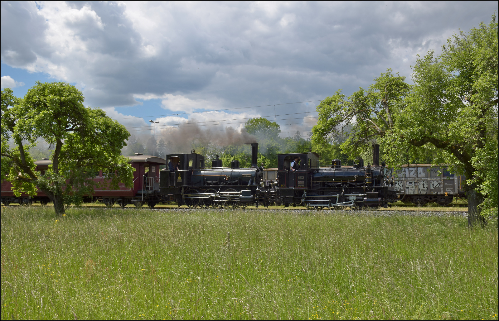 150 Jahre Emmentalbahn. 

E 3/3 853 der Jura-Simplon und Ed 3/3 3 'Langnau' der Emmentalbahn fahren mit ihrem Sonderzug durch einen Obstgarten in den Schwerindustriestandort Gerlafingen ein. Mai 2025.