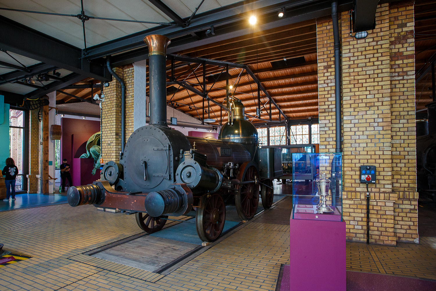 Der detailgetreue Nachbau von 1912 der Dampflok BEUTH, die erste eigenst�ndig in Deutschland entwickelte Dampflokomotive, ausgestellt im Deutschen Technikmuseum Berlin (bis 1996 Museum f�r Verkehr und Technik), hier am 22 September 2026.

Die von August Borsig 1844 konstruierte und in seine Borsigwerke in Berlin mit Werknummer 24 gebaute Lokomotive BEUTH gilt als die erste eigenst�ndig in Deutschland entwickelte Dampflokomotive. Vorher baute Borsig Lokomotiven nach amerikanischen Vorbildern nach. Die Lok gewann ein Wettrennen gegen ein Modell von Stephenson mit etwa zehn Minuten Vorsprung und galt f�r die folgenden zehn Jahre als Prototyp schneller deutscher Lokomotivkonstruktionen. Eine angetriebene Achse und zwei Laufachsen (1A1) sowie ein Stehkessel sorgten f�r vergleichsweise hohe Geschwindigkeiten (von bis zu 42 km/h. Sie bekam ihren Namen nach dem Leiter der preu�ischen Gewerbeakademie Christian Peter Wilhelm Beuth, der August Borsig prophezeit hatte, dass aus ihm nie etwas werden w�rde. Ein detailgetreuer Nachbau der Lok aus dem Jahr 1912 ist heute im Deutschen Technikmuseum Berlin ausgestellt, die Original-Lokomotive wurde 1864 ausgemustert und verschrottet.

Die Adler, von der englischen Lokomotivfabrik Robert Stephenson & Co., war 1835 die erste funktionsf�hige in Deutschland eingesetzte Dampflokomotive. Bereits 5 Jahre sp�ter (1840) baute Borsig die ersten Lokomotiven nach amerikanischen Vorbildern und ab 1844 nach eigenen Konstruktionen. 

Konstruktionsmerkmale
Die BEUTH besitzt gegen�ber dem ADLER au�enliegende, gr��ere Zylinder. Damit konnte auf eine aufw�ndig zu fertigende Kropfachse verzichtet werden. Die Treibstangen der Zylinder wirken direkt auf den in der Mitte liegenden Treibradsatz �ber exzentrisch in die Radsterne eingepresste Zapfen. Damit sind die Treibzapfen und Treibstangenlager mit h�chsten Scherkr�ften auch zum Abschmieren und zur Kontrolle leichter zug�nglich. Der h�here Dampfverbrauch des Antriebs erforderte einen leistungsst�rkeren und gr��eren Dampfkessel in Form eines Lang- und eines Stehkessels mit hochliegendem Dampfdom. Dieser kuppelartige Stehkessel ist typisch f�r alle fr�hen Borsig-Konstruktionen wie die Borsig und die auf die Beuth folgende Borussia. Der hintere Laufradsatz unter dem offenen F�hrerstand der Beuth kommt bei vielen sp�teren Konstruktionen ebenfalls vor. Die Beuth ist damit eigentlich die erste in Serie produzierte Dampflokomotive mit klassischen Merkmalen, wie sie weltweit in fast allen sp�teren Konstruktionen Verwendung fanden.

Fertigung und Einsatz
Die Beuth wurde 1844 auf der Berliner Gewerbeausstellung gezeigt und dann von der Berlin-Anhaltischen Eisenbahn eingesetzt. Diese Bauart wurde in insgesamt 71 Exemplaren bereits in einer Art Serie hergestellt und fand im gesamten norddeutschen Raum Verwendung. Die Weiterentwicklung der Beuth von Borsig ging als Borussia von 1844 z. B. auf der K�ln-Mindener Eisenbahn in Betrieb.

TECHNISCHE DATEN (Original):
Baujahr: 1844 
Ausmusterung: 1864
Hersteller: BORSIG GmbH in Berlin
Spurweite: 1.435 mm (Normalspur)
Achsfolge: 1A1 
L�nge �ber Puffer (Lok und Tender): 11.530 mm
Achsabstand: 3.810
Treibraddurchmesser: 1.524 mm
Laufraddurchmesser vorn/hinten: 990 mm
H�chstgeschwindigkeit: 42 km/h
Dienstgewicht: 14,4 t
Leistung: 68 PS PS (50 kW)
Zylinderanzahl: 2 (� 330 mm x 559 mm Hub)
Kessel�berdruck: 5,5 bar 
Rostfl�che: 0,83 m�
Verdampfungsheizfl�che: 46,56 m�
Gesamtheizfl�che: 46,6 m�
