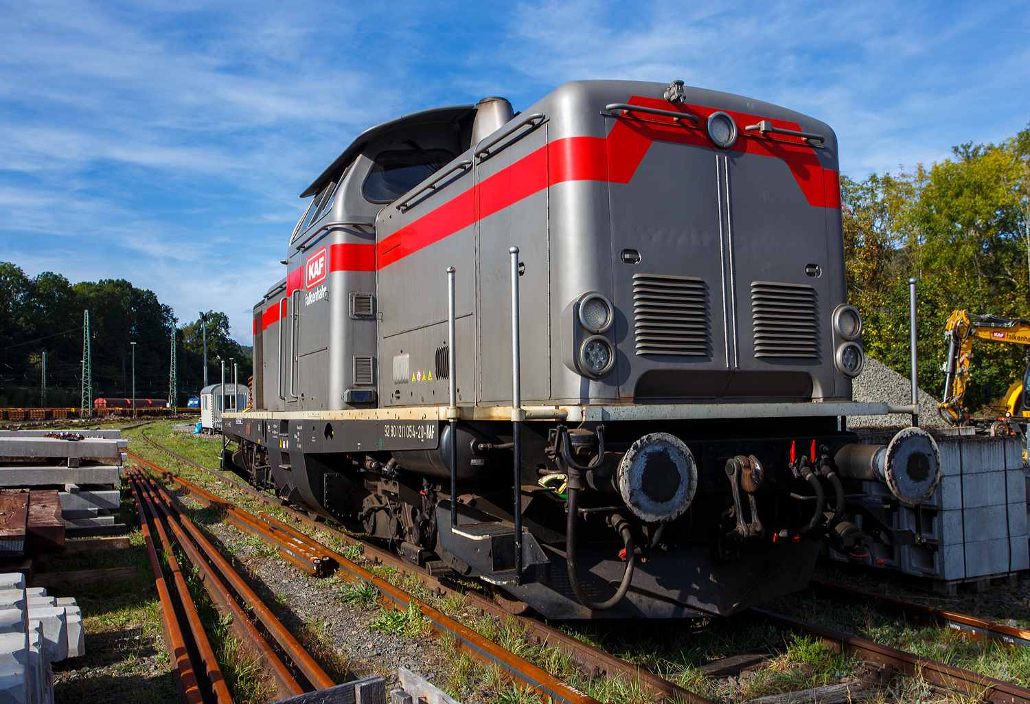 Die 211 054-2 (92 80 1211 054-2 D-KAF) der KAF Falkenhahn Bau AG (Kreuztal) ist am 03 Oktober 2026 beim Rbf Betzdorf/Sieg abgestellt. 

Die Streckendiesellokomotive V 100.10 wurde 1962 von MaK (Maschinenbau Kiel AG) in Kiel-Friedrichsort unter der Fabriknummer 1000072 gebaut und als V 100 1054 an die Deutsche Bundesbahn geliefert. Zum 01.01.1968 erfolgte dann die Umzeichnung in DB 211 054-2, als diese fuhr sie bis zur Ausmusterung 1999. Zum 15.06.1999 ging sie an den Eschenauer Kulturlokschuppen  Seku  e. V., im M�rz 2012 ging sie an ELV - Eisenbahn Logistik Vienenburg Willrich & M�hlberg GbR, und im Juni 2022 an die VEB - Vulkan-Eifel-Bahn Betriebsgesellschaft mbH in Gerolstein. Seit dem 23.09.2023 ist sie nun bei der KAF Falkenhahn Bau AG in Kreuztal.

Die V100.10 (BR 211) haben eine12-Zylinder-Viertakt-Dieselmotor mit etwas geringerer Leistung gegen�ber der V 100.20. Da die V 100.10 (BR 211) f�r den leichten Reise- und G�terzugdienst auf Nebenstrecken vorgesehen war. Bei den V 100.10 kam �berwiegend der Maybach MD 650 1B und der Daimler-Benz MB 820 Bb zum Einsatz, seltener war der MAN L 12 V 18/21 zu finden. Die Nennleistung lag bei 809 kW (1100 PS). Ab den 1980er-Jahren wurden die V 100.10 vermehrt mit st�rkeren MB 835 Ab-Motoren mit 990 kW (1.350 PS) ausger�stet (min. 135 Loks), so auch bei dieser Lok. Der Motor wurde jedoch auf 919 kW (1.250 PS) gedrosselt, damit das urspr�ngliche Str�mungsgetriebe mit seiner niedrigeren Getriebeeingangsleistung beibehalten werden konnte.

Erste Wahl f�r eine Lokomotive in der Leistungsklasse bis 1.100 PS w�re das bew�hrte hydraulische Dreiwandler-Getriebe LT306r aus der V 200.0 gewesen. Dieses Getriebe entsprach allerdings nicht mehr dem neuesten technischen Stand und bot keine Reserven zur Steigerung der Eingangsleistung �ber 1.100 PS hinaus. Die DB lie� deshalb von Voith ein neues Getriebe L216rs in Wandler-Wandler-Kupplung-Ausf�hrung entwickeln. Durch Verringerung der �bersetzung zwischen Getriebeeingangswelle und Hohlwelle lie� sich das Getriebe an h�here Motorleistungen anpassen. Dies wurde sp�ter f�r die Baureihe 212 genutzt. Das Getriebe ist als Einwellen-Getriebe konstruiert, das hei�t die Pumpenr�der laufen auf einer Hohlwelle, in der die Sekund�rwelle mit den Turbinenr�dern liegt. Die Leitr�der sind feststehend mit dem Getriebegeh�use verbunden. An den hydraulischen Teil schlie�t sich das mechanische Nachschaltgetriebe an, bestehend aus Wende- und Stufenschaltung. 

TECHNISCHE DATEN:
Spurweite: 1.435 mm (Normalspur)
Achsfolge: B'B'
Eigengewicht: 63,0 t
L�nge �ber Puffer: 12.300 mm
Drehzapfenabstand: 6.000 mm
Achsstand im Drehgestell: 2.200 mm
gr��te Breite: 3.115 mm
gr��te H�he �ber Schienenoberkante: 4.275 mm
kleinster befahrbarer Gleisbogen: 100 m
Treibraddurchmesser: 950 mm (neu)
H�chstgeschwindigkeit: 100 km/h
Dieselmotor: V-12-Zylinder-Dieselmotor mit Abgasturbolader Daimler-Benz MB 835 Ab (mit BBC VTR 250-Abgasturbolader), gedrosselt.
Hubraum: 78,25 Liter (Zylinderbohrung � 190 mm x 230 Hub)
Leistung: 919 kW (1.250 PS)
Nenndrehzahl: 1.500 U/min
Anfahrzugkraft: 177 kN
Getriebe: Voith L216rs
Kraft�bertragung: hydraulisch
Kupplungsart: Schraubenkupplung
