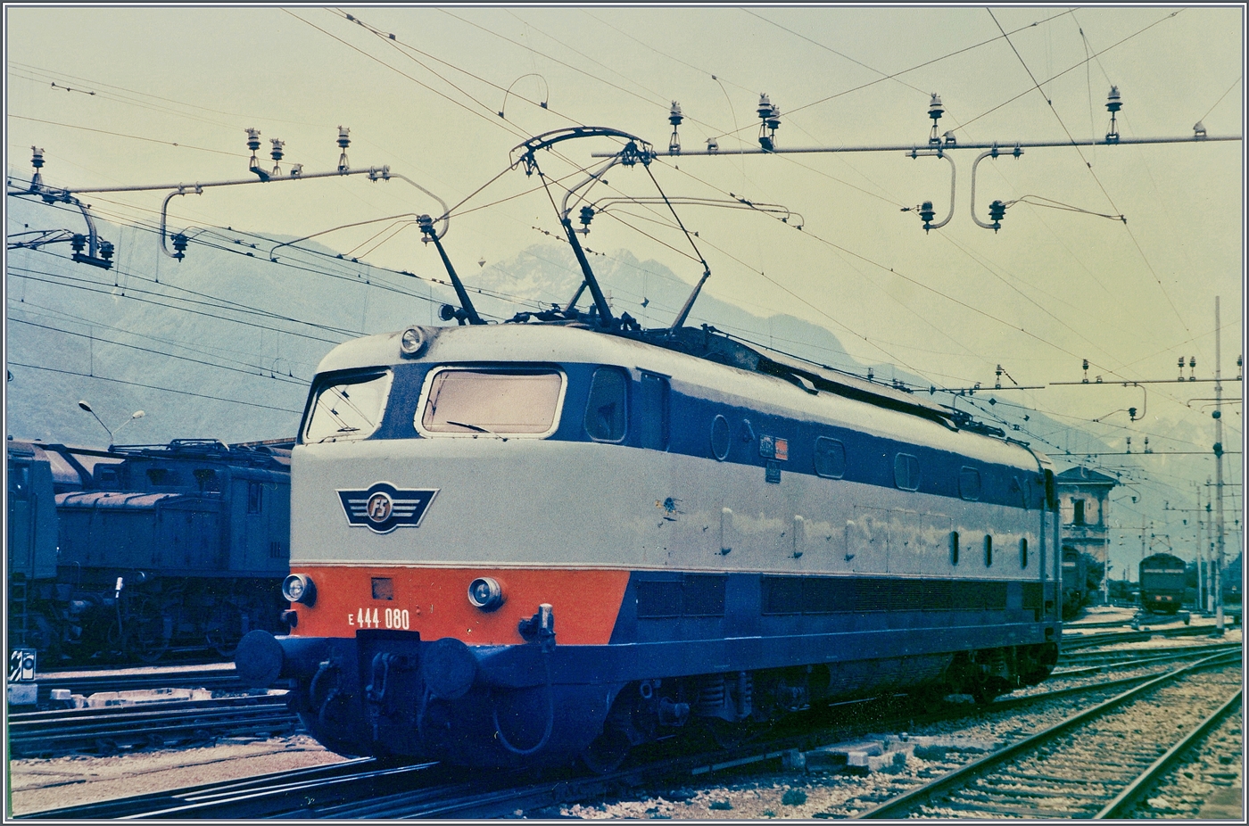 Die formsch�ne FS E 444 080  Tartaruga  (Schildkr�te) rangiert in Domodossola. Die FS verf�gte �ber 117 Loks dieser Baureihe, die mit einer maximalen Geschwindigkeit von 180 km/h im schnellen Schnellzugsverkehr zum Einsatz kamen, sp�ter aber im Frontbereich optisch wenig vorteilhaft umgebaut wurden aber noch lange Zeit im Einsatz standen. Neben der E 444 080 zeigen sich im Hintergrund einige FS E 626; die damals haupts�chlich im G�terverkehr eingesetzt wurden.

Analogbild vom Mai 1985 
