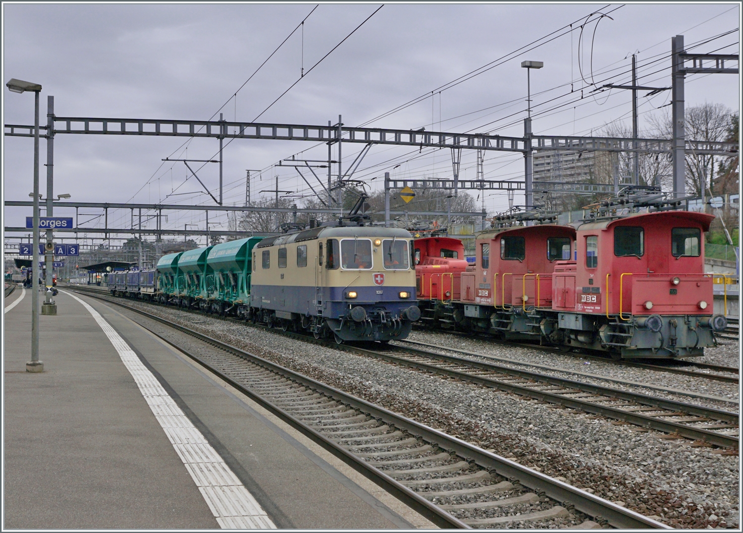 Die  IRSI Rheingold Re 4/4 II 11387 (Re 421 387) ist in Morges mit ihrem Kieszug von Gland angekommen und beginnt nun das Manöver, indem der Zug dann auf die Rollbockanlage geschoben wird. Neben der Lok stehen zwei BAM MBC Te 2/2 (Te 212) und (neu) der Tem III (97 85 1220 329-7. 

4. März 2024