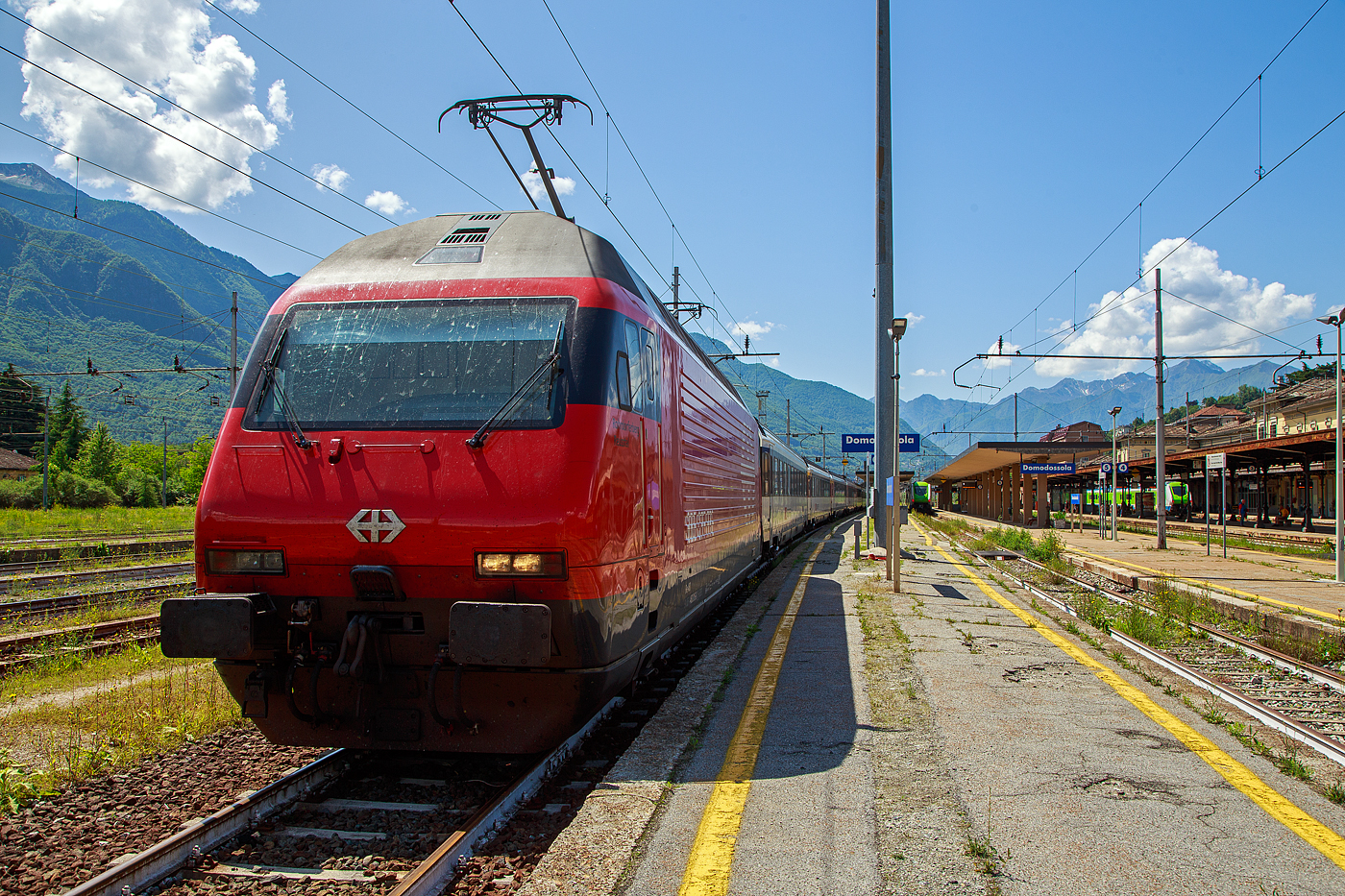 Die SBB Re 460 016-0  Rohrdorferberg Reusstal   (91 85 4 460 016-0 CH-SBB) hat am 26 Mai 2023 mit dem IR 3017 (Brig – Domodossola) den Zielbahnhof Domodossola (I) erreicht und ist nun mit dem Zug auf Gleis 6 abgestellt. Nach gut 2 ½ Stunden (um 13:48 Uhr) geht es dann als IR3022 von Domodossola zurück nach Brig.

Die Lok2000 wurde 1992 von der SLM (Schweizerischen Lokomotiv- und Maschinenfabrik in Winterthur) unter der Fabriknummer 5477 gebaut, der elektrische Teil ist von der ABB (Asea Brown Boveri). Sie war 2015 der 2. Prototyp der im SBB Werk Yverdon-les-Bains (VD) das vollständige Modernisierungs-/Refit-Programm LOK2000 durchlief. So sollen die Loks fit für die nächsten 20 Jahre sein, zudem werden durch alle 119 SBB Re 460 Lokomotiven so etwa 30 Gigawattstunden Energie eingespart.

Modernisierungs-/Refit-Programm der LOK2000 (Re 460):
Zwischen 2015 und November 2022 unterzog die SBB alle ihrer 119 Re 460 Lokomotiven (LOK2000) einem Refit-Programm (Modernisierung) im SBB Werk Yverdon-les-Bains (VD). Dabei wurden unter anderem die auf dem GTO-(Thyristor)-Halbleiter-Technologiestand der 1990er-Jahre basierende Leistungselektronik (GTO- Traktionsumrichter) durch IGBT-Traktionsumrichter (insulated-gate bipolar transistor) der neuesten Generation von ABB ersetzt, welche die Fahrmotoren mit Strom speisen und Bremsenergie zurückgewinnen. Die SBB hat damit, in Zusammenarbeit mit der ABB, die gesamte Flotte Re 460 modernisiert und hat diese auf den neuesten Stand der Antriebstechnik gebracht. Die gesamte Flotte spart dank neuen Traktionsumrichtern und weiteren technischen Optimierungen jedes Jahr rund 30 Gigawattstunden Energie ein, das sind fünf Gigawattstunden mehr als ursprünglich erwartet. Die Energieeinsparungen entsprechen dem durchschnittlichen Stromverbrauch von 10.000 Schweizer Haushalten. Die Umrüstung auf modernste IGBT-Technologie und mit Wasser statt Öl betriebene Kühlsysteme der Umrichter ermöglicht eine Senkung des Energieverbrauchs und sorgt für einen ökologischeren und sicheren Bahnbetrieb.

Geschichte und Beschreibung:
Die SBB bestellte im Zusammenhang mit den Projekten Bahn 2000 eine erste Serie von 12 Universal-Hochgeschwindigkeitslokomotiven, noch als Re 4/4VI – 10701 bis 10712 bestellt. Diese Bestellung wurde noch vor Beginn der Konstruktionsarbeiten auf 24 erhöht. Für den alpenquerenden Güterverkehr wurde von der Eidgenossenschaft eine weitere Serie von 75 Lokomotiven (auch als 'Hupac'-Lokomotiven bekannt) in Auftrag gegeben. Die vorgegebenen Liefertermine für dieses Los waren derart kurz, dass die SLM an die Kapazitätsgrenze ihrer Werkstätte gelangte. Es wurden deshalb dreißig Lokkasten an Krauss-Maffei in München untervergeben 

Im Hinblick auf die geplante Hochgeschwindigkeitsstrecke wurden nochmals zwanzig Lokomotiven nachbestellt, was die Serie auf 119 ansteigen ließ. Die im Volksmund Lok 2000 genannte Lokomotive hat eine Höchstgeschwindigkeit von 230 km/h und eine Leistung von 6,1 MW und ist für den Schnellzugs- wie Güterverkehr ausgelegt. Um die Gesamtmasse von 84 Tonnen nicht zu überschreiten musste ein gesickter Leichtbaukasten konstruiert werden, dessen Form vom Designstudio Pininfarina entworfen wurde.

Um auf den Strecken durch die Alpen den Radverschleiß gering zu halten, haben die Lokomotive radial einstellbare Achsen erhalten. Auch auf dem Gebiet der elektrischen Ausrüstung wurde Neuland beschritten. Zwei Ebenen Steuerelektronik für das Fahrzeug und den Antrieb sind eingebaut. Über das Fahrzeugleitgerät wurden zu viele Elemente gesteuert, wie Scheibenwischer oder Signalhorn. Dies führte anfänglich zu vielen Störungen und oft blieben Lokomotiven auf der Strecke liegen. Erst die Verbesserung der Programmierung erlaubte es die Zuverlässigkeit so zu erhöhen, dass es kaum mehr zu Ausfällen führte. Als Feststellbremse wurden in den Drehgestellen Permanentmagnetschienenbremsen eingebaut. Auch hier gab es am Anfang übermäßig viele Störungen und die Lokomotiven klebten regelmäßig an den Schienen fest.

Um den Schiebedienst auf den Gebirgsstrecken weniger personalaufwendig durchzuführen, wurden mehrere Lokomotiven mit einer amerikanischen Funkfernsteuerung ausgerüstet und als Ref 460 bezeichnet. Damit konnte von der Zugspitze aus eine am Zugschluss eingesetzte Lokomotive ferngesteuert werden. Die Divisionalisierung der SBB erlaubte es nicht mehr groß davon zu profitieren, denn alle Lokomotiven wurden dem Personenverkehr zugeteilt und der Einsatz vor Güterzügen war somit beendet.

Die Re 460 blieb nicht ein Einzelstück und entwickelte sich zu einer Lokomotivfamilie. Eine erste Bestellung wurde von der BLS für acht Lokomotiven gemacht. Die BLS Re 465 hat im Gegensatz zur Re 460 vier statt zwei Stromrichter. Jeder Motor hat dadurch seine eigene Regelung um die Eigenschaften im Bergeinsatz zu verbessern. Weiter können die BLS-Lokomotiven mit allen gängigen Schweizer Lokomotiven in Vielfachsteuerung verkehren. Das erlaubte es bei der BLS den Lokomotiveinsatz flexibler zu gestalten. Die SBB bestellte ebenfalls zehn Re 465, die bei Ablieferung schon das blaue BLS-Design hatten, aber erst später von der BLS übernommen wurden. Nach Finnland konnten auch in zwei Serien Lok 2000 verkauft werden. Dort sind sie als Sr2 bezeichnet. Nach Versuchsfahrten in Norwegen im schwierigen Winterdienst bestellten die NSB die fast baugleichen Lokomotive EL18 2241 bis 2262. Eine letzte Lieferung umfasst zwei Lokomotiven für Hongkong. Sie werden an jedem Ende eines Doppelstockzuges eingesetzt. Dies sind die letzten von SLM in Winterthur gebauten Fahrzeuge. Die Re 460 ist bis auf weiteres die letzte Vollbahn-Streckenlokomotive, die komplett von schweizerischen Unternehmen konstruiert und gebaut wurde. Der Lokomotivbau in der Schweiz wurde inzwischen aus wirtschaftlichen Gründen eingestellt. Einzig Stadler Rail stellt in der Schweiz noch Lokomotiven für den Rangier- und für den Zahnradbetrieb her.

TECHNISCHE DATEN der Re 460:
Nummerierung:  Re 460 000–118
Anzahl: 119
Baujahre: 1991–1996
Hersteller: SLM - Winterthur / ABB (Asea Brown Boveri)
Design: Pininfarina
Hersteller Lokkasten: teilweise (30 Stück) Krauss-Maffei (München)
Spurweite: 1.435 mm (Normalspur)
Achsformel: Bo'Bo'
Länge über Puffer: 18.500 mm
Höhe: 4.300 mm
Breite: 3.000 mm
Drehzapfenabstand: 11.000 mm
Achsabstand im Drehgestell:  2.800 mm
Laufraddurchmesser: 1.110 mm (neu)
Dienstgewicht: 84,0 t
Höchstgeschwindigkeit:  200 km/h (zugelassen)  / 230 km/h (Techn. möglich) 
Stundenleistung: 6.100 kW / 8.300 PS
Dauerleistung: 4.800 kW / 6.000 PS
Anfahrzugkraft: 300 kN
Dauerzugkraft: 275kN
Stromsystem: 15 kV, 16,7 Hz (über Oberleitung)