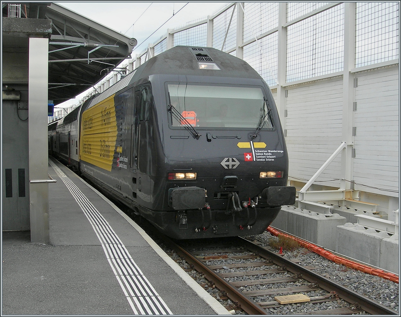 Die SBB Re 460 028 (Re 460 91 85 4 460 028-4 CH-SBB) tr�gt die gef�llige Werbedekoration  Schweizer Wanderwege  wobei die Lok wahrscheinlich mit der gelben Front noch besser wirkt. 
Die SBB Re 460 028 ist mit dem IR 95 1712 von Brig nach Gen�ve A�roport und konnte beim Halt in Lausanne fotografiert werden. 

4. Juni 2026