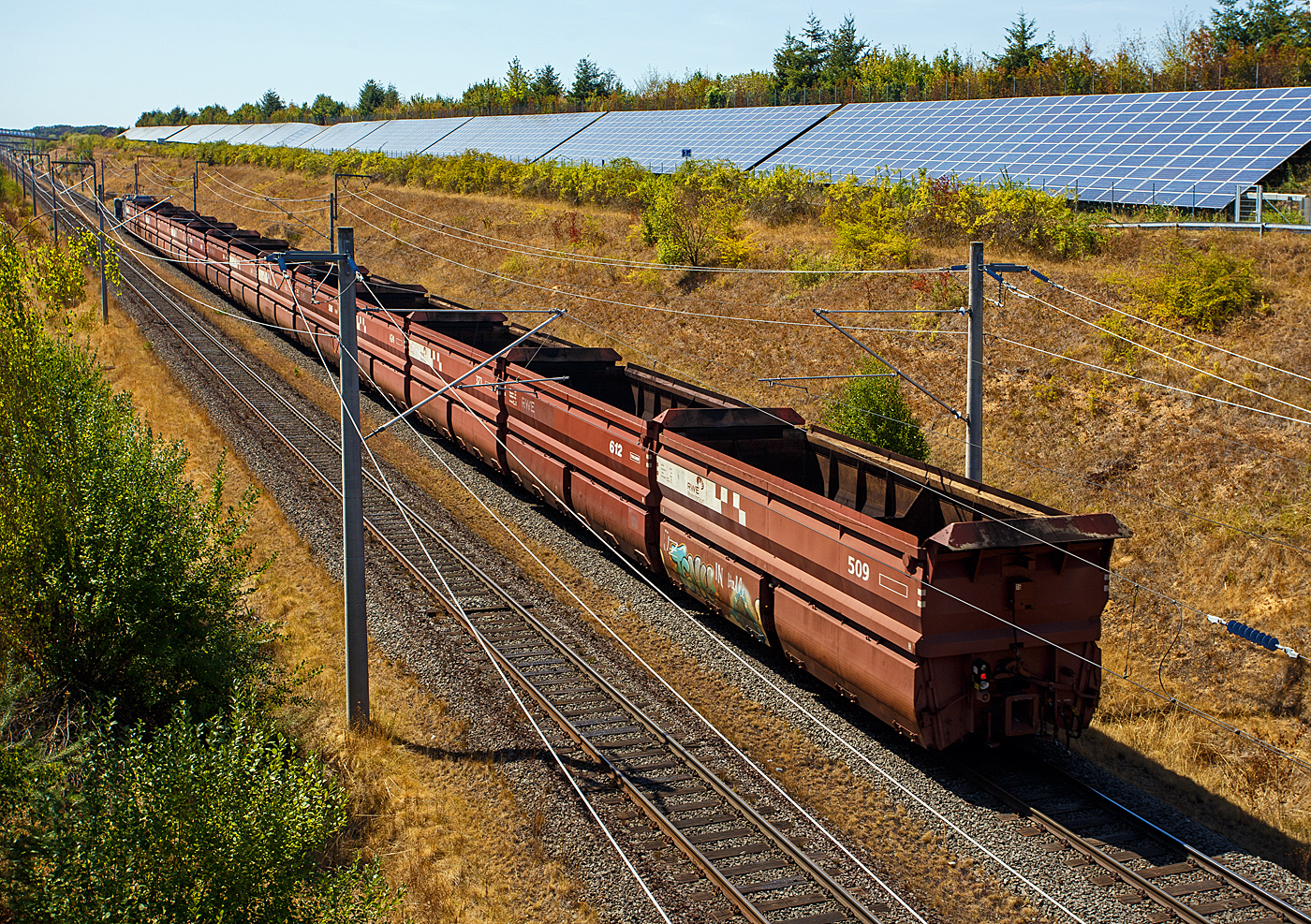Die Tagebaulokomotive El 1 – 549 der RWE Power AG (ex Rheinbraun) kommt am 12 August 2026 bei Kerpen-Buir auf der Hambachbahn mit einen Braunkohle Selbstentladewagen Leerzug, von den Kraftwerken zum Tagebau Hambach.

Die elektrischen Tagebaulokomotiven EL 1 wurden von einem Konsortium, bestehend aus den Firmen Henschel, Krupp, Krauss-Maffei, Siemens-Schuckertwerke, AEG und Brown, Boveri & Cie., in der Zeit von 1954 bis 1965 in 117 Exemplaren f�r die Rheinischen Braunkohlenwerke (sp�ter Rheinbraun, heute RWE Power AG) gefertigt. Die Lok 549 wurde 1956 von Krauss-Maffei in M�nchen-Allach unter der Fabriknummer 18209 gebaut, der elektrische Teil ist von AEG. Sie ist aktuell eine von noch 21 El 1 Loks im Einsatzbestand.

Die Hambachbahn ist eine Tagebau-Werksbahn der RWE Power (ex Rheinbraun) im Rheinischen Braunkohlerevier. Sie bindet den Tagebau Hambach an die Nord-S�d-Bahn an, �ber die die RWE-Kraftwerke in Niederau�em, Frimmersdorf und Neurath mit Braunkohle versorgt werden.

Die 1983 in Betrieb genommene Hambachbahn wird, wie auch die Nord-S�d-Bahn, elektrisch betrieben, jedoch als Besonderheit abweichend zum �blichen Bahnstrom mit Einphasenwechselstrom 6,6 kV und 50 Hz. 

Die Streckenl�nge in der Spurweite 1.435 mm (Normalspur), betr�gt 21,00 Kilometer. Die beginnt beim Kohlebunker Hambach (Verladestation) und endet beim Stellwerk Fn in Bergheim-Auenheim, wo sie dann an die Nord-S�d-Bahn anschlie�t. Im Bereich des Kohlenbunkers des Tagebaus Hambach befindet sich eine Wendeschleife.

Im Rahmen der Erweiterung des Tagebaus Hambach wurde der Streckenverlauf zusammen mit der Bundesautobahn 4 um etwa drei Kilometer nach S�den verlegt und beide verlaufen seit September 2014 parallel zur Schnellfahrstrecke K�ln–Aachen der Deutschen Bahn, es gibt jedoch keine direkte Verbindung zwischen den beiden Strecken. Die neue Strecke verl�uft aus Landschafts- und Schallschutzgr�nden in einem etwa 8 bis 15 Meter tiefen Einschnitt im Gel�nde. Aber im Gegensatz zur deutlich vernehmbaren DB Hauptstrecke und zum sehr lauten Rauschen der Autobahn n�hern sich die Braunkohlez�ge kaum h�rbar. 

