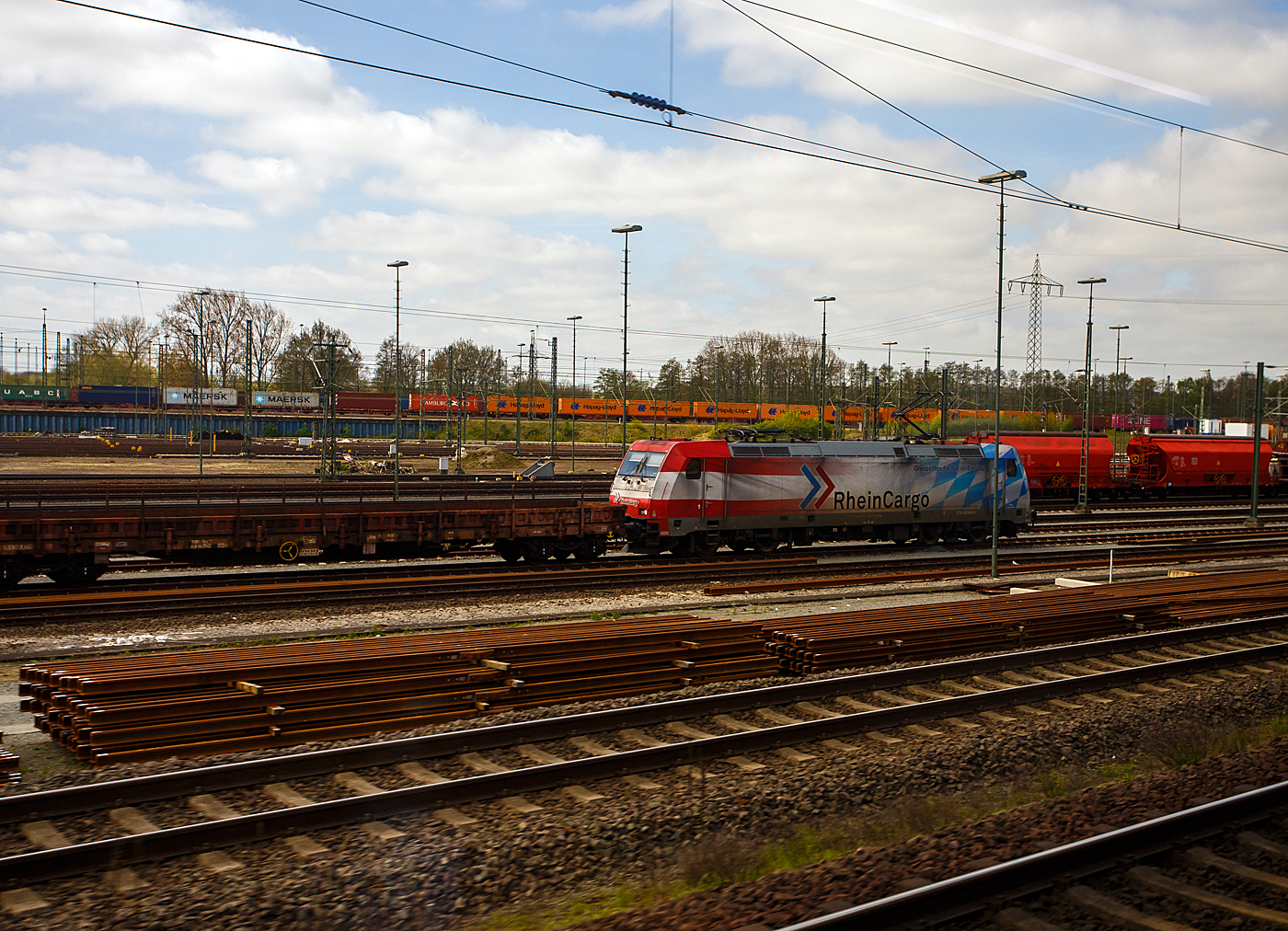 Ein Blick aus dem Zug auf den Rangierbahnhof Maschen (Maschen Rbf) am 23 April 2026, hier steht die 185 350-6 mit Werbung „Grenzenlos f�r Sie im Einsatz“ (91 80 6185 350-6 D-RHC), ex DB Cargo 185 350-6, der RheinCargo GmbH & Co. KG, mit einem Flachwagenzug. 

Die TRAXX F140 AC2 wurde 2008 von Bombardier in Kassel unter der Fabriknummer 34250 gebaut und die DB Cargo (damals Railion) geliefert, 2019 wurde sie an die RheinCargo GmbH & Co. KG verkauft. 

Maschen Rangierbahnhof, s�dlich von Hamburg an der Bahnstrecke Hannover–Hamburg ist der gr��te Rangierbahnhof Europas und nach dem Rangierbahnhof Bailey Yard im US-Bundesstaat Nebraska der zweitgr��te Rangierbahnhof der Welt. Der Rangierbahnhof nimmt bei einer L�nge von 7 km und einer maximalen Breite von 700 Metern eine Fl�che von 280 Hektar (2,8 km�) ein. Bei der Er�ffnung betrug die Gesamtgleisl�nge 300 Kilometer. Als zweiseitiger Rangierbahnhof verf�gt Maschen �ber zwei Zugbildungsanlagen. Im Nord-S�d-System wurden eine Richtungsgruppe mit 48 Gleisen und eine Einfahrgruppe mit 16 Gleisen erbaut, das S�d-Nord-System bekam eine Richtungsgruppe mit 64 Gleisen und eine Einfahrtsgruppe mit 17 Gleisen. Beide Systeme werden durch Abstell- und Ordnungsgruppen erg�nzt. Zudem sind eine achtgleisige Wagenausbesserungshalle zur Reparatur von G�terwagen und ein Bahnbetriebswerk zur Instandhaltung von elektrischen und dieselgetriebenen G�terzuglokomotiven mit einer zweigleisigen Untersuchungshalle und zahlreichen Abstellgleisen im Freien vorhanden. F�r jedes System sind zwei Stellwerke zust�ndig.