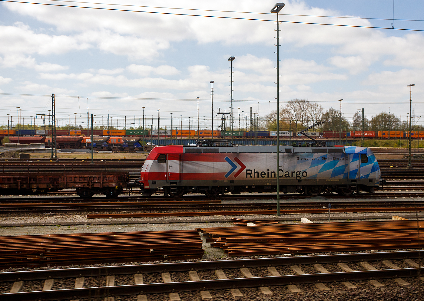 Ein Blick aus dem Zug auf den Rangierbahnhof Maschen (Maschen Rbf) am 23 April 2026, hier steht die 185 350-6 mit Werbung „Grenzenlos f�r Sie im Einsatz“ (91 80 6185 350-6 D-RHC), ex DB Cargo 185 350-6, der RheinCargo GmbH & Co. KG, mit einem Flachwagenzug. 

Die TRAXX F140 AC2 wurde 2008 von Bombardier in Kassel unter der Fabriknummer 34250 gebaut und die DB Cargo (damals Railion) geliefert, 2019 wurde sie an die RheinCargo GmbH & Co. KG verkauft. 

Maschen Rangierbahnhof, s�dlich von Hamburg an der Bahnstrecke Hannover–Hamburg ist der gr��te Rangierbahnhof Europas und nach dem Rangierbahnhof Bailey Yard im US-Bundesstaat Nebraska der zweitgr��te Rangierbahnhof der Welt. Der Rangierbahnhof nimmt bei einer L�nge von 7 km und einer maximalen Breite von 700 Metern eine Fl�che von 280 Hektar (2,8 km�) ein. Bei der Er�ffnung betrug die Gesamtgleisl�nge 300 Kilometer. Als zweiseitiger Rangierbahnhof verf�gt Maschen �ber zwei Zugbildungsanlagen. Im Nord-S�d-System wurden eine Richtungsgruppe mit 48 Gleisen und eine Einfahrgruppe mit 16 Gleisen erbaut, das S�d-Nord-System bekam eine Richtungsgruppe mit 64 Gleisen und eine Einfahrtsgruppe mit 17 Gleisen. Beide Systeme werden durch Abstell- und Ordnungsgruppen erg�nzt. Zudem sind eine achtgleisige Wagenausbesserungshalle zur Reparatur von G�terwagen und ein Bahnbetriebswerk zur Instandhaltung von elektrischen und dieselgetriebenen G�terzuglokomotiven mit einer zweigleisigen Untersuchungshalle und zahlreichen Abstellgleisen im Freien vorhanden. F�r jedes System sind zwei Stellwerke zust�ndig.
