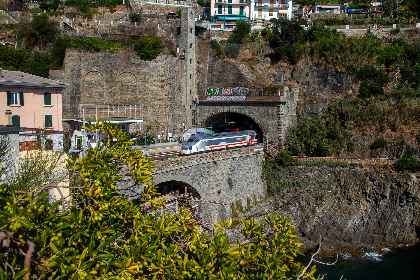 Ein Trenitalia Intercity (IC) rauscht im Sandwich zweier E.414 (ehemalige Triebköpfe E.404 A der ersten ETR 500 – Monotensione) durch den Cinque Terre Bahnhof Riomaggiore in Richtung Genua (Genova).

Vor dem Zuganfang auf der anderen Bahnsteigseite sieht am den Eingang von dem Fußgänger-Tunnel vom Bahnhof zum Zentrum von Riomaggiore. Dieser führt durch den Eisenbahntunnel, ist aber abgegrenzt. 

Zudem sieht man hinten auch den hohen Lift (Aufzug), mit diesem kommt man auf den etliche Meter oben liegenden Weg. Der Lift ist aber in der Saison kostenpflichtig, aber er erspart etliche Treppenstufen oder einen gewaltigen Umweg.