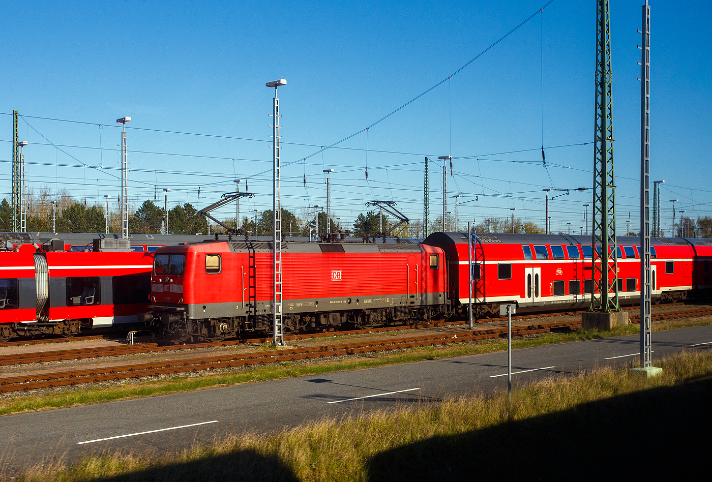 Eine Wende-Lok, die 112 119-3 (91 80 6112 119-3 D-DB) der DB Regio Nordost, ex DR - Deutsche Reichsbahn 112 119-3 steht 22 April 2026 mit einem DoSto-Zug in Rostock in der Abstellgruppe. Aufnahme aus einem Zug heraus. 

Die 112 wurde zu einem Symbol f�r die Deutsche Einheit, denn sie war die erste Lokomotivbaureihe, die von beiden Bahnverwaltungen gemeinsam beschafft wurde. Man kam �berein, dass DR und DB jeweils 45 Loks der leicht verbesserten Baureihe 112.1 bei AEG Schienenfahrzeuge Hennigsdorf GmbH bestellen w�rden. Die AEG hatte 1992 zwischenzeitlich ihr 1946 enteignetes Werk in Hennigsdorf wieder �bernommen, das zu DDR-Zeiten VEB Kombinat Lokomotivbau Elektrotechnische Werke „Hans Beimler“ Hennigsdorf (LEW) hie�.

So wurde die 112 119-3 von AEG 1993 unter der Fabriknummer 21514 gebaut und an die DR geliefert. Mit der Bahnreform und Fusion der beiden deutschen Staatsbahnen (Deutschen Bundesbahn und der Deutschen Reichsbahn), wurde die Lok zum 01.01.194 zur 112 119-3 der DB AG. 