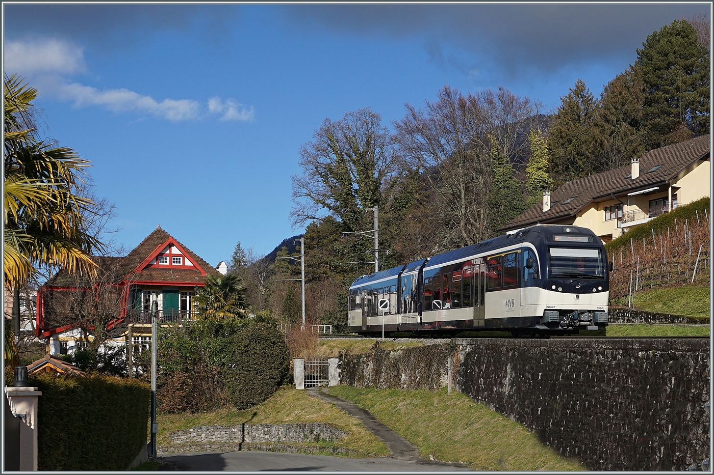 Kaum 19 Minuten nach der Abfahrt im verschneiten Les Avants erreicht der CEV MVR ABeh 2/6 7504  Vevey , der als R 34 2317 nach Montreux unterwegs ist, den Halt Planchamp (530 müM). Wie man unschwer feststellen kann ist der Zug nun in einer grünen Landschaft unterwegs, wobei bei genauerem Hinsehen sogar Plamen die Strecke säumen.

3. Januar 2025