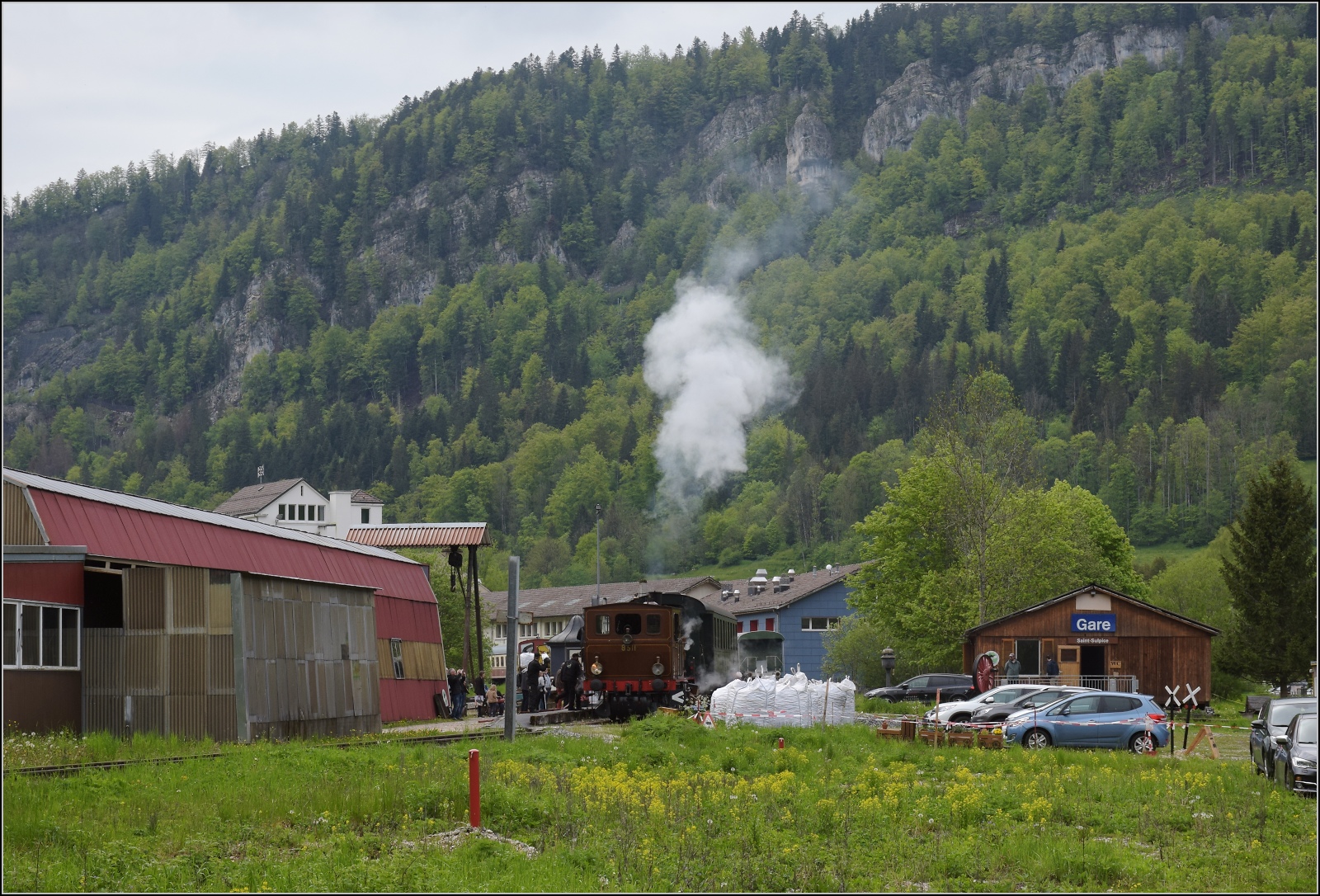 Vapeur Val-de-Travers: Train du Terroir.

Bereitstellung des Zuges von und mit E 3/3 8511 in St-Sulpice. 

Die etwas unmotivierte Motivauswahl gilt es allerdings zu erklären. Wer genau ins Bild schaut, erkennt etwas links der Dampfwolke unterhalb der Unterbrechung der Felswand zwei Masten und ganz links im Bild auch noch mal. Die Hauptstrecke nach Frankreich via dem zuvor gezeigten Bahnhof Les Verrières geht durch diese Felswand ins noch einmal 200 m höher gelegene Hochtal zur Wasserscheide. St-Sulpice an der Karstquelle der Areuse und später Buttes mussten daher regional erschlossen werden. Das wurde seinerzeit durch die RVT gemacht, die heute in die TransN integriert ist. Das schaffte immerhin dieses kleine Biotop für die Dampfenthusiasten des VVT. Dazu Mai 2023. 