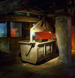 Auch eine Eisenbahn (Schwebebahn): Die H�ngebahn der V�lklinger H�tte am 01 Juli 2025, hier einer der selbstfahrenden H�ngebahnwagen unter der M�llerhalle. 

Die H�ngebahn ist eine Einschienenbahn bzw. Schwebebahn mit selbstfahrende H�ngebahnwagen, bei den Schr�gaufz�gen auf die Gichtb�hne wurden sie mit Seilz�gen unterst�tzt. 

Die H�ngebahn war seit 1911 in Betrieb. 1985 fuhren pro Stunde 240 beladene H�ngebahnwagen zur  Gicht . Sie liefen �ber die sogenannte Gichtb�hne in einer H�he von 27 Metern, die Rohstoffe (Eisenerz, Sinter, Schrott, Kalk und Koks) um die 6 Hoch�fen von dort zu beschicken. Die Gleisl�nge der H�ngebahn betrug �ber 6 km.

Der M�ller (Eisenerz, Sinter, Schrott und Kalk wurden in der M�llerhalle (s�d�stlich der Hoch�fen) in selbstfahrende H�ngebahnwagen verladen und �ber einen vierspurigen Schr�gaufzug auf die Gichtb�hne gezogen. Auf der Gichtb�hne laufen mehrere H�ngebahnspuren an den sechs Hoch�fen vorbei und wieder zur�ck zum Schr�gaufzug, mit kreisf�rmigen Abzweigschleifen um jeden Hochofen herum. Auf der Nordwestseite der Gichtb�hne l�uft ein kleinerer zweispuriger Schr�gaufzug hinunter zur Kokerei, um den Koks f�r die Hoch�fen zu f�rdern.

Die Gichtb�hne verbindet alle sechs Hoch�fen mit einer Ebene. Auf dieser Ebene werden die H�ngebahnwagen entlanggef�hrt. Die Gichtb�hne ist ca. 240 Meter lang und erstreckt sich in einer H�he von exakt 27 Meter.  Die Gichtb�hne hat Ihren Namen von dem Wort  Begichten , was das Bef�llen der Hoch�fen mit den Rohstoffen f�r die Eisenerzeugung bezeichnet. Eisenerz, Sinter, Zusatzstoffe, Koks, aber auch eisenhaltige Reststoffe aus der Industrie oder Schrott wurden in die Gicht, den Einf�lltrichter des Hochofens, gekippt. Die „Haube/obere Glocke“, der obere Verschluss des Hochofens, wurde angehoben, dann f�nf H�ngebahnwagen um den Trichter gefahren. Unterhalb der Haube befindet sich ein zweiter Verschluss, die „Gicht-Glocke“. Sie blieb geschlossen, um das Gichtgas aufzufangen. Das Material wurde auf die Glocke gekippt, dann die Haube abgesenkt. Erst wenn die Haube geschlossen war, wurde die Glocke nach unten gefahren: Das Material rutschte in den Hochofenschacht. Diese Verschlussart nannte man „Doppelglockenverschluss“, bei modernen Hoch�fen werden heute glockenlose Gichtverschl�sse verwendet. 
